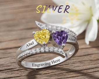 Könnte beinhalten: Ein silberner Ring mit zwei herzförmigen Edelsteinen, einem gelben und einem lila, besetzt mit kleinen, klaren Steinen. Der Ring ist mit den Namen "Abel" und "Alice" sowie den Worten "Engraving Here" graviert. Das Wort "Silver" ist ebenfalls sichtbar.