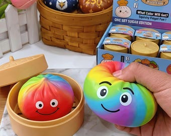 Könnte beinhalten: Bunte, quetschbare Zappelspielzeuge in Form von Brötchen. Eines ist regenbogenfarben und wird in einer Hand gehalten, ein anderes ist rot mit einem lächelnden Gesicht. Die Verpackung trägt die Aufschrift "BUN" und "Squishy Fidget Fun!"