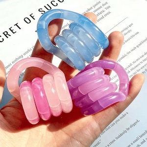 May include: Three colorful, translucent hand-shaped rings in shades of blue, pink, and purple. The rings are made of a smooth, glossy material and are held in a hand. The text "CRET OF SUC" is visible in the background.