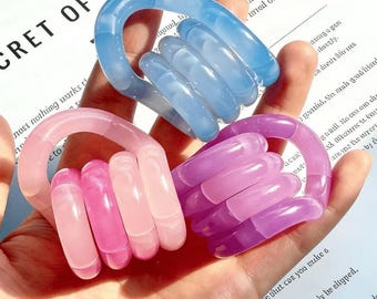 Könnte beinhalten: Drei durchscheinende, handförmige Ringe in Blau, Rosa und Lila. Die Ringe bestehen aus einem glatten, glänzenden Material. Die Ringe werden in einer Hand gehalten. Der Text "CRET OF SUC" ist im Hintergrund sichtbar.