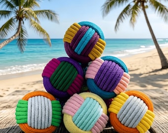Könnte beinhalten: Eine Sammlung farbenfroher, gewebter Bälle in verschiedenen Blau-, Lila-, Rosa-, Gelb- und Orangetönen. Die Bälle sind auf einer Holzoberfläche mit einem Strand und Palmen im Hintergrund angeordnet. Die Bälle bestehen aus dickem, geflochtenem Seil.