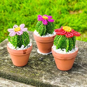 May include: Three small decorative cactus figures crafted from green beads, each potted in a terracotta-coloured clay pot filled with white gravel. Each cactus features a beaded flower in light pink, bright magenta, or vibrant red with yellow centres.