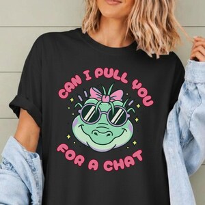 May include: A black short-sleeved t-shirt featuring a graphic of a light green alligator wearing pink sunglasses and a pink bow. The text "CAN I PULL YOU FOR A CHAT" is printed in pink letters in a circular arrangement around the alligator illustration.