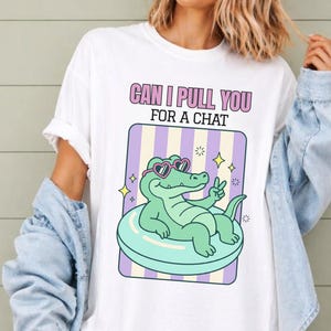 May include: A white t-shirt featuring a graphic of a green alligator wearing heart-shaped sunglasses, lounging on a light blue pool float. The text above reads CAN I PULL YOU FOR A CHAT, set against a background of vertical purple and cream stripes.