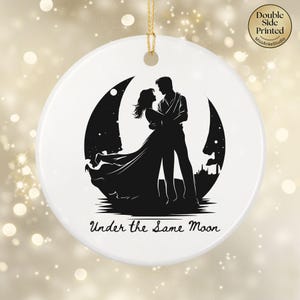 May include: A round white ceramic ornament featuring a black silhouette of a couple embracing in front of a large crescent moon. The text "Under the Same Moon" is printed in black script below the figures. A gold string is attached to the top.