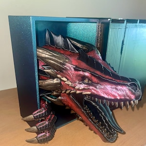 May include: A detailed sculpture of a dragon's head and claws emerging from a blue box. The dragon features red, black, and gold coloring, with sharp teeth and textured scales. The box has a glittery finish, suggesting a fantasy or decorative theme.