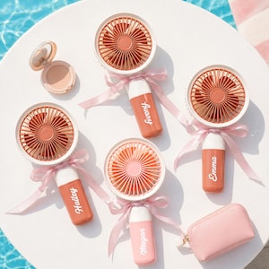 Puede incluir: Varios ventiladores de mano blancos y coral, cada uno adornado con una cinta rosa y un nombre personalizado. También hay un estuche de maquillaje compacto abierto y una pequeña bolsa rosa sobre una superficie blanca.