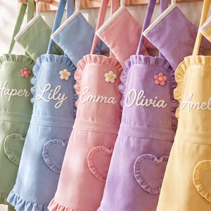 May include: Five corduroy aprons in sage green, light blue, pink, lavender, and yellow, each featuring a ruffled trim, a heart-shaped pocket, a small flower embellishment, and a white embroidered name: Haper, Lily, Emma, Olivia, and Amelia.