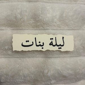 May include: A rectangular off-white paper label with torn, deckled edges featuring the black Arabic text "ليلة بنات" printed in the centre. The label has a smooth, matte texture and is positioned horizontally against a soft, white, fuzzy surface.