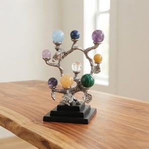 Pode incluir: Uma escultura de árvore em metal prateado com ramos e folhas texturizados, montada sobre uma base quadrada preta. A escultura sustenta nove pedras preciosas esféricas polidas de várias cores, incluindo azul claro, azul escuro, roxo, amarelo, transparente e verde.