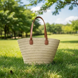 Peut inclure: Un sac fourre-tout en paille tressée beige avec des poignées et des accents en cuir marron. Le sac est posé sur de l'herbe verte dans un parc, avec des arbres et du soleil en arrière-plan. Le sac a une large ouverture et une forme structurée.
