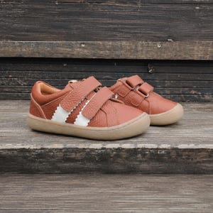 Może przedstawiać: Para niskich sneakersów z teksturowanej brązowej skóry. Każdy but posiada dwa paski zapinane na rzepy oraz dwa pionowe białe paski z boku. Sneakersy mają zaokrąglony nosek i płaską, jasnobrązową gumową podeszwę.