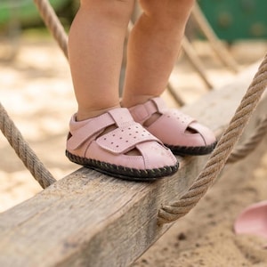 May include: A pair of pink baby sandals with a black sole. The sandals have a strap closure and small perforations on the top. The sandals are on a wooden beam with a rope on either side.