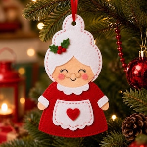 May include: A felt Christmas ornament shaped like a grandmother figure with white hair, a red dress, and a white apron featuring a red heart. The ornament has a scalloped collar, pink cheeks, and a small green holly sprig with red berries on the hair.