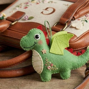 May include: A green felt dragon ornament with red accents, wings, and a smiling face. The dragon has a star and floral embroidery on its side. A brown leather bag with floral embroidery is in the background.