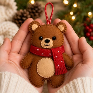 May include: A brown felt bear ornament featuring a tan belly and inner ears, wearing a red scarf with white cross-stitch detailing and a red and white patterned hanging loop. The bear has black bead eyes and a stitched nose and mouth.