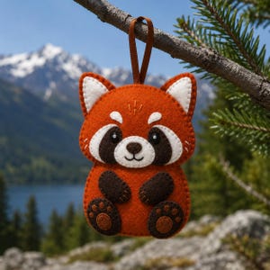 May include: A felt red panda ornament featuring a vibrant orange body, white ear tips, and dark brown eye patches. The creature has a small stitched nose and mouth, dark brown paws, and a brown ribbon loop at the top for hanging purposes.