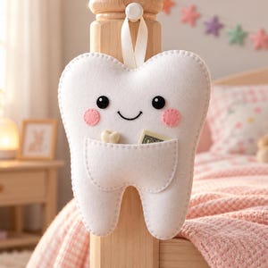 May include: A white felt tooth-shaped pouch with a smiling face, black eyes, and pink cheeks. The pouch features a small pocket containing a miniature tooth and a folded banknote, suspended by a white ribbon from a wooden bed post.