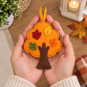 May include: A felt tree ornament featuring a rounded orange canopy with a dark brown trunk. The canopy is decorated with four small felt leaves in red, yellow, green, and orange, each with stitched vein details. A yellow ribbon loop is attached to the top.