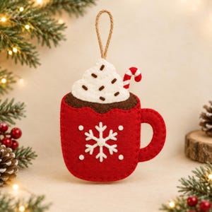 May include: A red felt Christmas ornament shaped like a mug of hot cocoa with white whipped cream, brown sprinkles, and a red and white candy cane. A white snowflake is centered on the mug, which hangs from a tan twine loop.