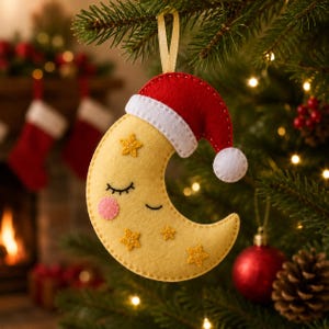 Könnte beinhalten: Ein Filzanhänger in Form einer Mondsichel mit einer roten Weihnachtsmütze, die einen weißen Rand und einen weißen Bommel hat. Der gelbe Mond zeigt ein schlafendes Gesicht mit geschlossenen Augen, rosa Wangen und vier kleinen gelben Sternen.
