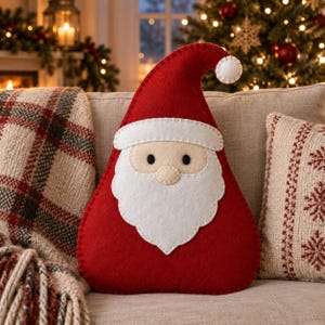 Könnte beinhalten: Ein rotes Filzkissen in Form eines Weihnachtsmanns mit weißem Bart, weißem Schnurrbart, weißem Mützenrand und einem weißen Bommel an der Mützenspitze. Das Gesicht hat zwei kleine schwarze runde Augen und eine beige Nase mit sichtbaren Nähten.
