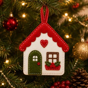 May include: A felt house-shaped Christmas ornament featuring a white facade, a scalloped red roof, and a red hanging ribbon. The design includes a green arched door, a brown window with a red flower box, and a small red heart centred above.