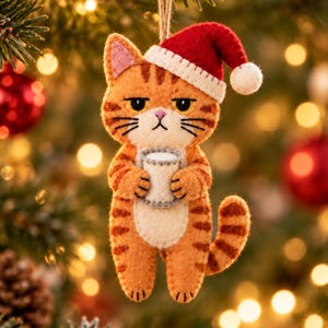 Può includere: Un ornamento in feltro a forma di gatto tigrato arancione con strisce scure, che indossa un cappello da Babbo Natale rosso e bianco. Il gatto ha un'espressione imbronciata e tiene una piccola tazza bianca con entrambe le zampe.