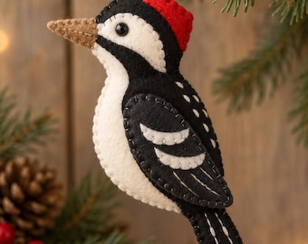 Puede incluir: Un adorno de fieltro con forma de pájaro carpintero en colores negro, blanco y rojo con un pico color canela. El ave está confeccionada con costuras visibles en los bordes e incluye un pequeño lazo de cuerda para colgarlo.