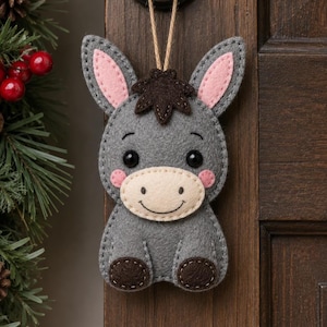 May include: A gray felt donkey ornament featuring a cream-colored muzzle, dark brown hair tuft and hooves, pink inner ears, and pink cheeks. The donkey has black bead eyes and a stitched smile, suspended by a thin tan string for hanging purposes.