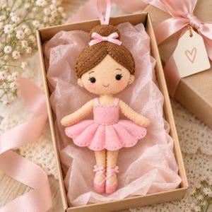 May include: A felt ballerina doll ornament with brown hair in a bun, a pink bow, and a pink tutu. The doll features stitched facial details, pink ballet slippers with ribbons, and a hanging loop, presented inside a gift box with pink tissue paper.