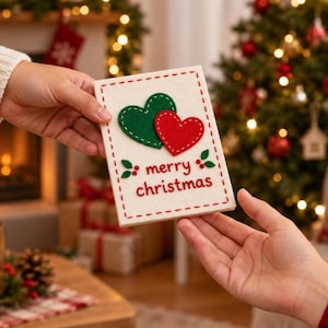 May include: A rectangular white felt card featuring a large green heart and a smaller red heart stitched onto the front. The text merry christmas is embroidered in red below the hearts, surrounded by a red stitched border and small green leaves.
