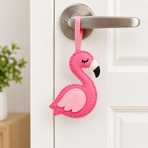 May include: A pink felt flamingo ornament hanging from a silver door handle by a matching pink ribbon. The flamingo features a lighter pink wing, a black stitched eye, and a black-tipped beak, with visible stitching along the edges of the felt.