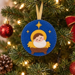 May include: A circular felt Christmas ornament featuring a blue background with a yellow star and smaller stars. The center depicts a brown manger with a swaddled infant wearing a yellow halo. The ornament hangs from a gold ribbon loop.