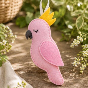 May include: A pink felt parrot ornament with a yellow crest and a black beak. The bird has a white ribbon for hanging and is decorated with stitched details. The ornament is designed to look like a cockatoo.