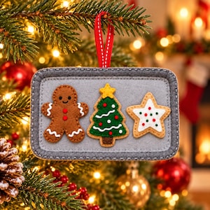 May include: A rectangular gray felt ornament featuring three stitched appliqués: a brown gingerbread person, a green Christmas tree with a yellow star, and a white star-shaped cookie with red and green dots, hanging from a red ribbon.