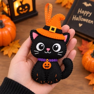 Pode incluir: Um enfeite de feltro preto em forma de gato com um chapéu de bruxa laranja, faixa preta e fivela amarela, uma coleira roxa com um pingente de abóbora laranja e uma fita branca com bolinhas para pendurar, tudo com costuras visíveis.