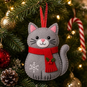 Puede incluir: Un adorno navideño de fieltro gris con forma de gato que lleva una bufanda roja con hojas de acebo verdes y bayas rojas, un copo de nieve blanco bordado, orejas rosas y una cinta roja para colgarlo en el árbol de Navidad.