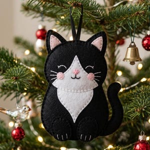 May include: A black and white felt cat ornament with pink accents, hanging on a Christmas tree. The cat has a white chest, pink cheeks, and a smiling face. Other ornaments and lights are visible.