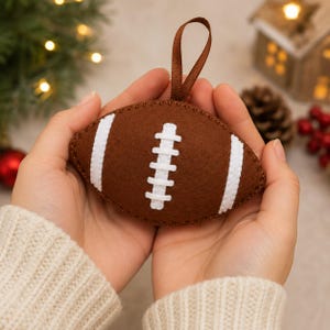 May include: A brown felt football ornament featuring white stitched laces and two white stripes on each side. The football has a brown ribbon loop attached to the top for hanging, with visible stitching along the edges of the felt material.