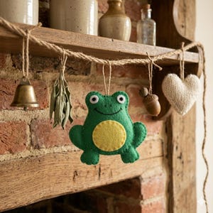 May include: A green felt frog ornament with a yellow belly and white eyes hangs from a jute rope. Other hanging decorations include a brass bell, dried leaves, acorns, and a knitted heart. The decorations are displayed on a wooden shelf against a brick wall.