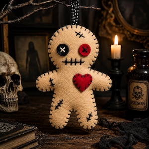 Op de afbeelding: Een beige vilt voodoo-pop ornament met een zwart knoop-oog, een rood knoop-oog en een gestikte mond. De pop heeft een rood vilten hart op de borst, zwarte festonsteken langs de randen en decoratieve zwarte kruissteekaccenten.