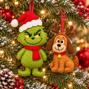May include: Two felt Christmas ornaments hanging from a tree. One is a green character with a red scarf and a red Santa hat with white trim. The other is a brown dog with reindeer antlers, a red collar, and a small yellow bell charm.