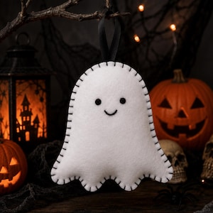 May include: A white felt ghost ornament featuring a simple embroidered black face with two circular eyes and a curved mouth. The ghost has a scalloped bottom edge and is outlined with black blanket stitching, suspended by a black satin ribbon loop.