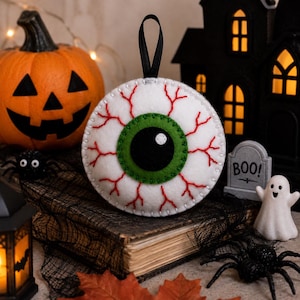 May include: A round felt ornament shaped like a human eyeball with a white sclera, a green iris, and a black pupil. The white surface features red embroidered veins and a black ribbon loop at the top for hanging. The edges are stitched with white thread.