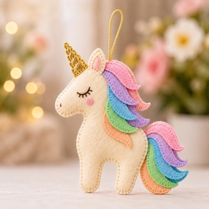 Puede incluir: Un adorno de unicornio de fieltro color crema con un cuerno de purpurina dorada, ojos cerrados y una mejilla rosa. La crin y la cola están compuestas por tiras de fieltro superpuestas en tonos pastel rosa, morado, azul, verde y naranja.