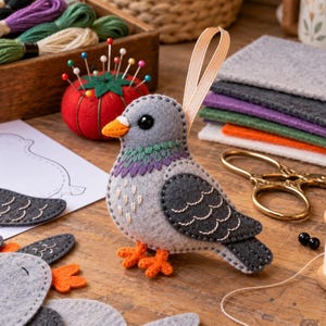 Peut inclure: Un ornement de pigeon en feutrine fait main avec un corps gris clair, des ailes gris foncé à coutures blanches, un bec et des pattes orange, et un col violet et vert. L'oiseau possède une boucle en ruban beige et un œil en perle noire.