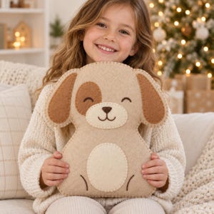 Puede incluir: Un peluche de fieltro con forma de perro en color tostado y marrón con cara cosida, que presenta una nariz marrón oscuro, una boca sonriente y ojos cerrados. El juguete tiene una mancha en el vientre de color crema y orejas marrones.