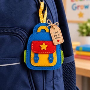 May include: A felt backpack-shaped keychain in blue, yellow, and red, featuring a yellow star on the front pocket. A tan tag attached with twine reads First day of school with a small red heart. The keychain hangs from a yellow polka-dot ribbon.