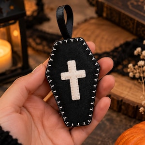 May include: A black felt coffin-shaped ornament featuring a white cross appliqué in the centre and white whip-stitched edges. A black ribbon loop is attached to the top for hanging. The item is constructed from dark fabric with contrasting stitching.